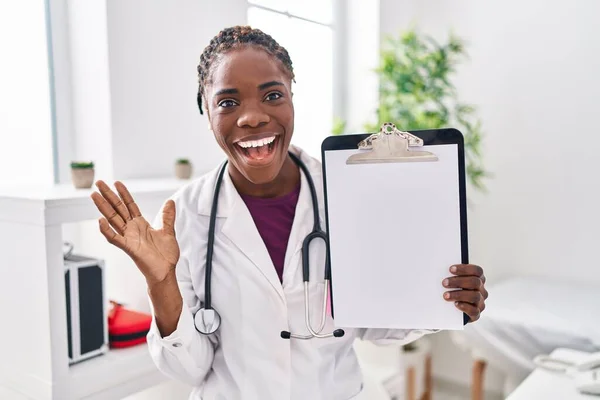幸せな笑顔で勝利を祝うクリップボードを保持し 手を上げて勝者の式を持つ美しい黒の医師の女性 — ストック写真