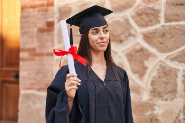 Young hispanic woman wearing graduated uniform holding diploma at university