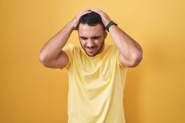 Young hispanic man standing over yellow background suffering from headache desperate and stressed because pain and migraine. hands on head. 