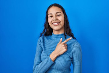 Genç Brezilyalı bir kadın mavi izole edilmiş arka planda neşeli, yüzünde bir gülümsemeyle mutlu ve doğal bir ifadeyle elini ve parmağını kenarı işaret ediyor. 