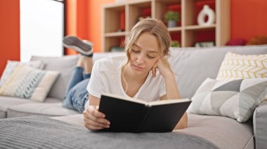 Young blonde woman reading book lying on sofa at home