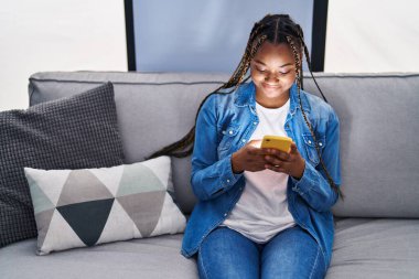 Evdeki koltukta oturan Afro-Amerikan kadın akıllı telefon kullanıyor