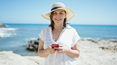 Orta yaşlı İspanyol turist kumsalda akıllı telefon kullanarak gülümsüyor.