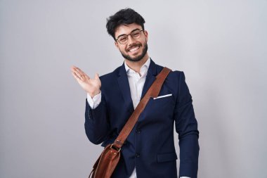 Sakallı, iş kıyafetleri giymiş, neşeli bir şekilde gülümseyen, avuç içiyle kameraya bakan İspanyol bir adam.. 