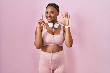 Spor kıyafetleri ve kulaklıkları takan Afrikalı Amerikalı kadın altı numaralı parmaklarını gösterip gösterirken kendinden emin ve mutlu bir şekilde gülümsüyor.. 