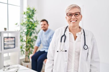 Tıbbi klinikte üniforma ve steteskop giyen kadın doktor dişlerini gösteren kendine güvenen bir gülümsemeyle ayakta ve mutlu görünüyor. 