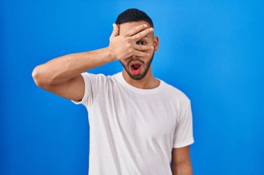 Young hispanic man standing over blue background peeking in shock covering face and eyes with hand, looking through fingers with embarrassed expression. 