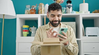 Genç İspanyol adam yemek odasında karton kutuları açmak için akıllı telefon kullanıyor.
