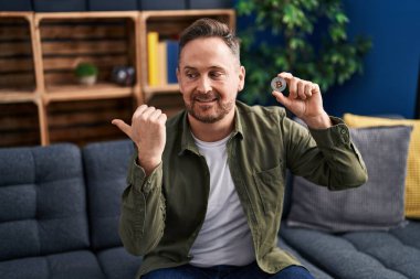 Orta yaşlı beyaz adam elinde sanal para tutuyordu. Bitcoin parmağıyla yan tarafı gösteriyordu. Ağzı açık gülümsüyordu. 