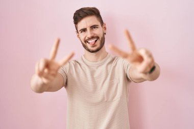 Pembe arka planda sakallı, ağzı açık bir şekilde gülümseyen ve parmaklarını zafer işareti yapan İspanyol bir adam. İki numara.. 