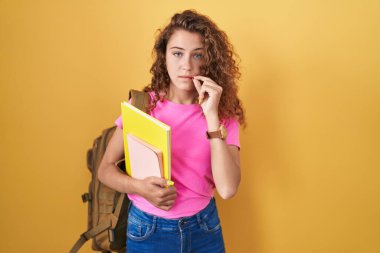 Young caucasian woman wearing student backpack and holding books mouth and lips shut as zip with fingers. secret and silent, taboo talking 