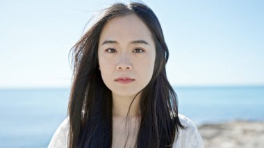 Genç Çinli kadın deniz kenarında ciddi bir ifadeyle ayakta duruyor.