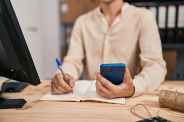 Young caucasian man ecommerce business worker using smartphone writing on notebook at office