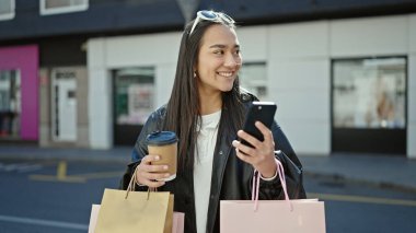 Genç ve güzel İspanyol kadın akıllı telefon kullanıyor. Elinde alışveriş poşetleri ve sokakta kahve tutuyor.