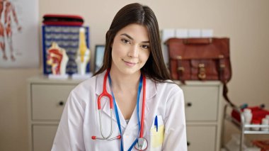 Young beautiful hispanic woman doctor smiling confident sitting on table at clinic