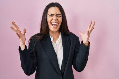 Genç esmer kadın pembe arka planda iş stili giyiniyor deli gibi bağırıyor ve agresif bir ifade ve kollarla bağırıyor. hüsran kavramı. 
