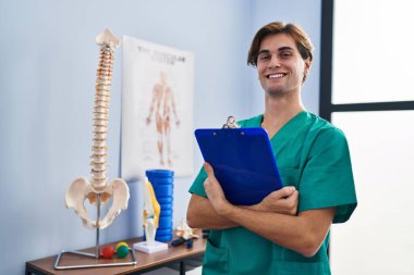 Young caucasian man pysiotherapist smiling confident holding clipboard at rehab clinic