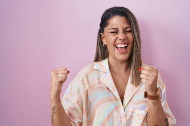 Blonde woman standing over pink background very happy and excited doing winner gesture with arms raised, smiling and screaming for success. celebration concept. 