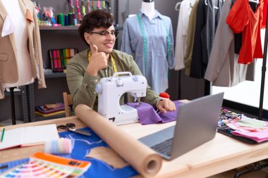 Genç, ikili olmayan terzi dizüstü bilgisayarlı, mutlu ve olumlu bir şekilde gülümseyen, mükemmel ve onay işareti yapan bir tasarımcı. 