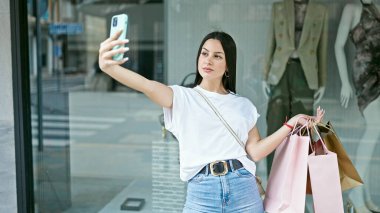 Genç ve güzel İspanyol bir kadın alışverişe çıkıyor. Elinde çantalarla elbise mağazasında akıllı telefonlardan selfie çekiyor.