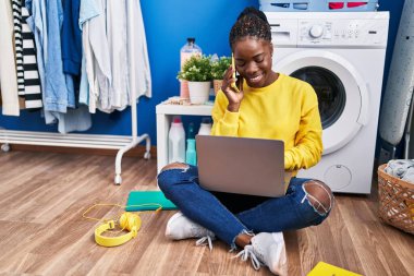 Akıllı telefon başında konuşan Afrikalı Amerikalı kadın çamaşır odasında çamaşır makinesini bekliyor.