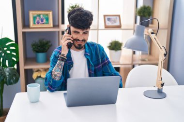 Genç İspanyol adam dizüstü bilgisayar kullanıyor ve evde akıllı telefonla konuşuyor.