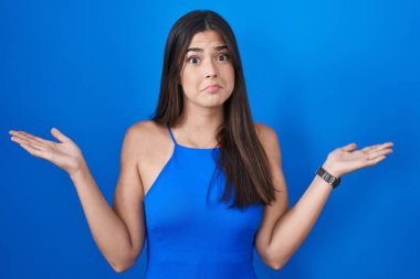 Hispanic woman standing over blue background clueless and confused expression with arms and hands raised. doubt concept. 