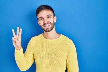 Hispanic man standing over blue background showing and pointing up with fingers number three while smiling confident and happy. 