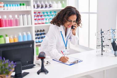 Eczanedeki dokümanlarda akıllı telefondan konuşan orta yaşlı kadın eczacı.