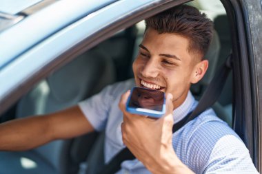 Young hispanic man talking on the smartphone sitting on car at street