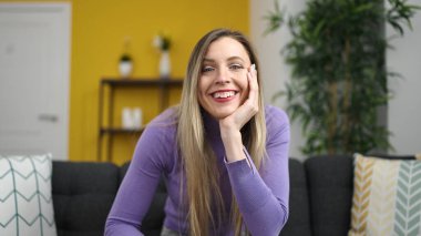 Sarışın genç bir kadın evdeki koltukta kendinden emin bir şekilde oturarak gülümsüyor.