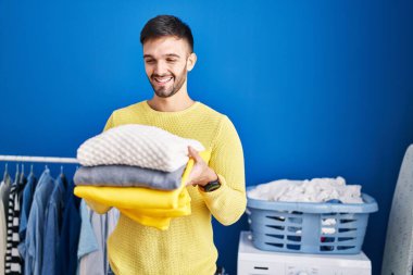 Çamaşırhanede katlanmış giysiler tutan kendine güvenen İspanyol genç adam.