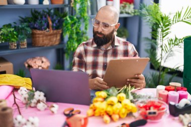 Young bald man florist using laptop reading document at flower shop