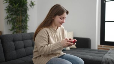 Young blonde woman playing video game sitting on sofa at home