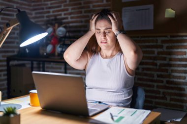 Geceleri ofiste çalışan esmer kadın baş ağrısı çekiyor. Acı ve migren yüzünden çaresiz ve stresli. Eller başının üstünde.. 