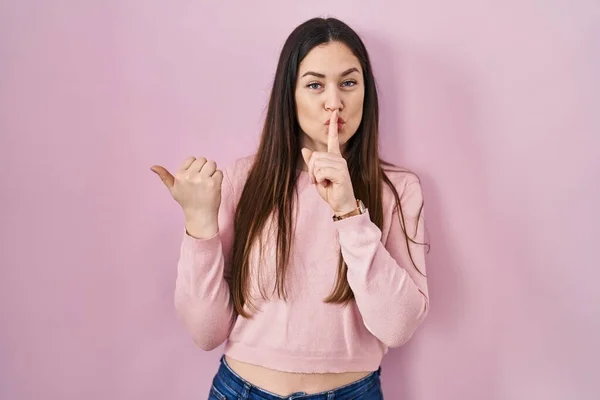 若いブルネットの女性がピンクの背景の上に立って 唇に手で側に指して指で静かになるように求めている 沈黙と秘密の概念 — ストック写真