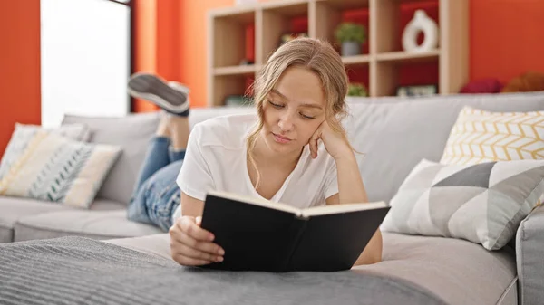 Young Blonde Woman Reading Book Lying Sofa Home — Zdjęcie stockowe