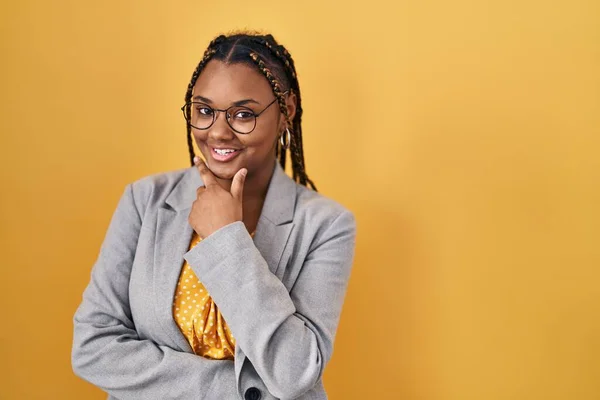 African american woman with braids standing over yellow background looking confident at the camera smiling with crossed arms and hand raised on chin. thinking positive.