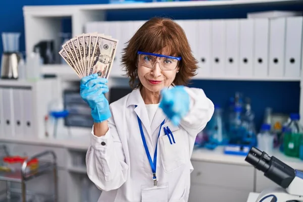 Middle age woman working at scientist laboratory holding money pointing with finger to the camera and to you, confident gesture looking serious