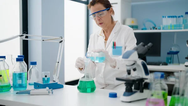 stock image Middle age hispanic woman wearing scientist uniform pouring liquid on test tube at laboratory