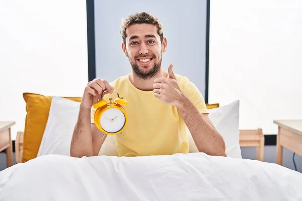 ベッドの中で目覚まし時計を保持髭を持つヒスパニック系の男は幸せと肯定的な笑みを浮かべて 親指アップ素晴らしいと承認サインを行う — ストック写真