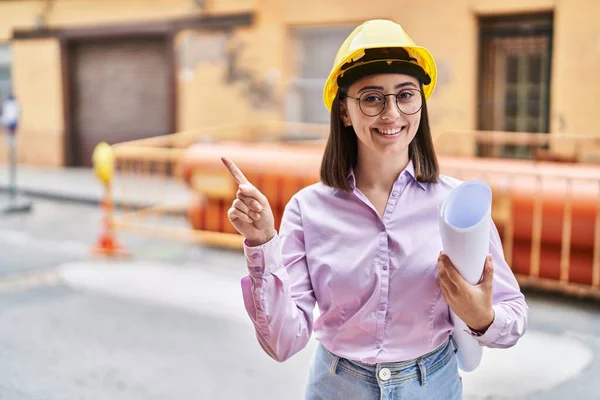建筑工地上戴着建筑师硬礼帽的西班牙裔女孩高兴地微笑着 手指手画脚地指向旁边 — 图库照片