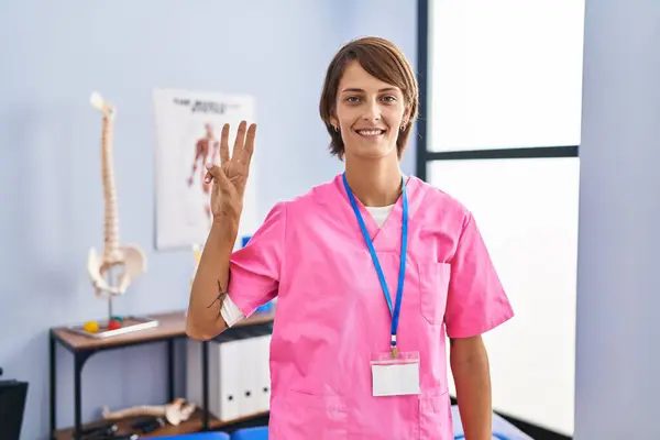 在康复诊所工作的布鲁内特妇女露出并用第3号手指指指指点点 同时带着自信和快乐的微笑 — 图库照片