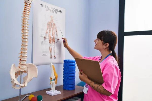 Młoda Piękna Latynoska Fizjoterapeutka Czytająca Dokument Patrzący Model Anatomiczny Klinice — Zdjęcie stockowe
