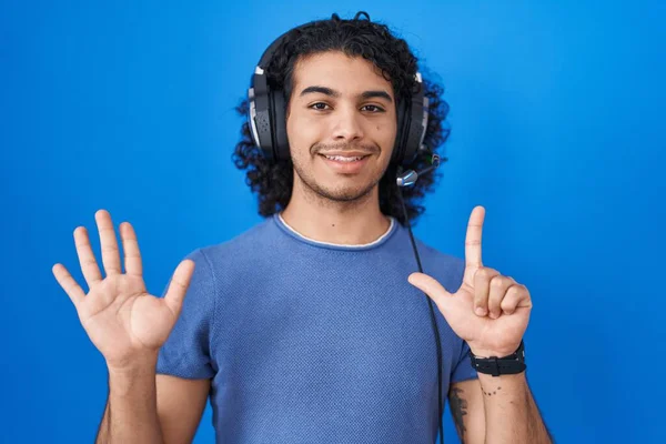 头发卷曲的西班牙裔男人一边用耳机听音乐 一边用7号手指指尖 同时带着自信和快乐的笑容 — 图库照片