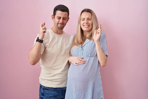 Casal Jovem Esperando Bebê Sobre Fundo Rosa Gesto Dedo Cruzado — Fotografia de Stock