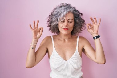 Middle age woman with grey hair standing over pink background relaxed and smiling with eyes closed doing meditation gesture with fingers. yoga concept. 