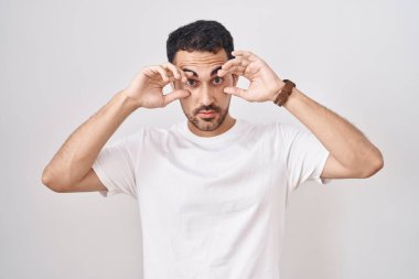 Beyaz arka plan üzerinde duran yakışıklı İspanyol adam parmaklarıyla gözlerini açmaya çalışıyor, uykulu ve yorgun sabah yorgunluğu için. 