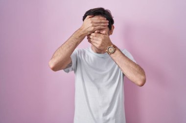 Young hispanic man standing over pink background covering eyes and mouth with hands, surprised and shocked. hiding emotion 