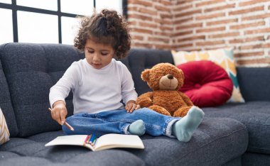 Adorable hispanic girl drawing on notebook sitting on sofa at home